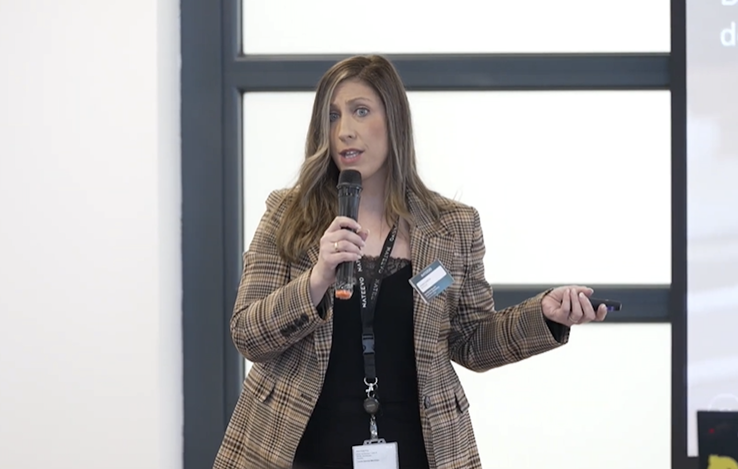 Una mujer con el pelo largo y castaño, vestida con una americana de cuadros escoceses y un top negro, sostiene un micrófono y gesticula mientras habla sobre marketing en tiempo real en un almuerzo de NATEEVO, con su cordón y su tarjeta de identificación visibles delante del fondo de la ventana.